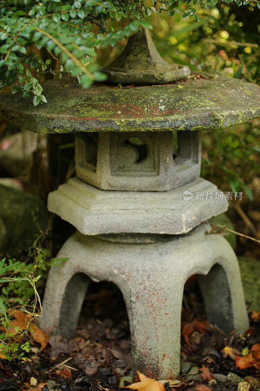 大型花岗岩kinitu rokkaku yukimi石雪灯的形象，在东方日本花园的竹叶背景和高大的现代方形柱灯的背景，风化的苔藓和地衣生长在表面，禅宗灯笼